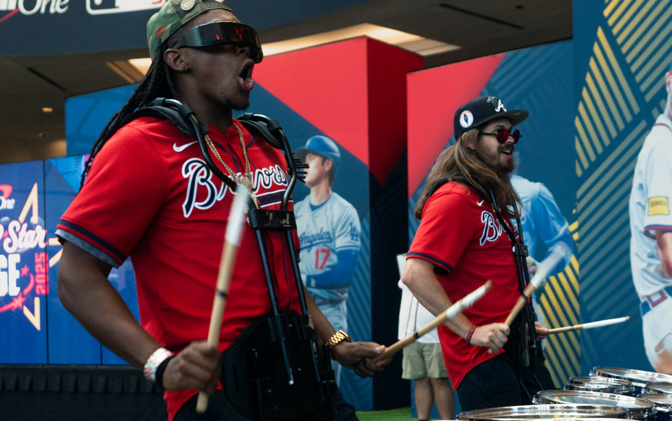 Live drumline performance in Atlanta at MLB All-Star Village 2025 with performers in Braves gear playing drums beneath large event banner